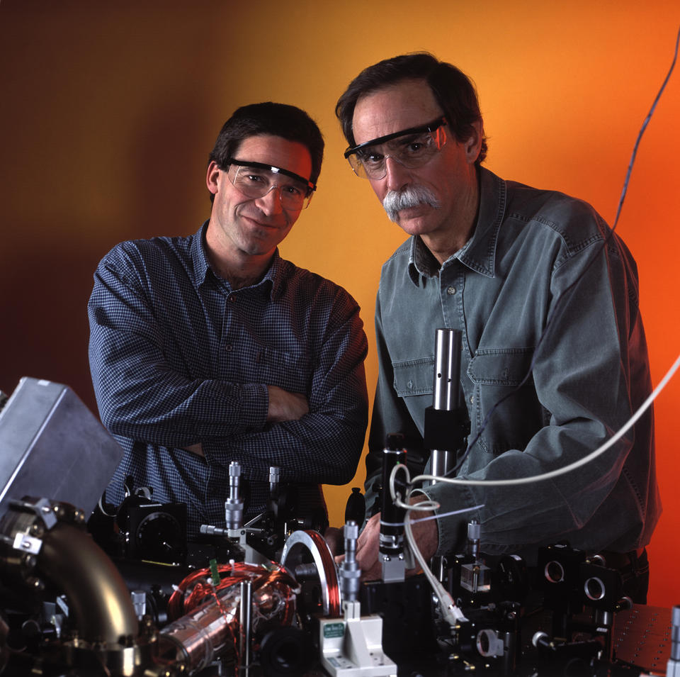 David Wineland and Dietrich Leibfried wear goggles and stand in front of equipment