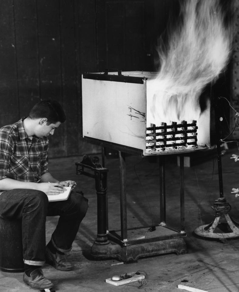 Black and white photo of a young man hunched over sitting on a chair next to a large white box that is burning a wood fire for an experiment.