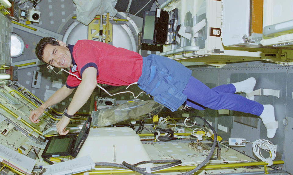 Color photograph of man wearing red shirt and bluejeans floating inside space shuttle. The space shuttle silver walls are covered in yellow pipes and red buttons from floor to ceiling.