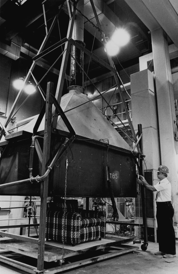 Black and white photo of a man standing in front of large piece of metal equipment shaped like a funnel. It reaches from the floor to the ceiling.