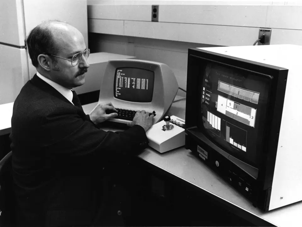 Black and white photo of man working on two computer screens in 1989. The screen to the left is small and long and the screen to the right is large and boxy, both show correlating data. 