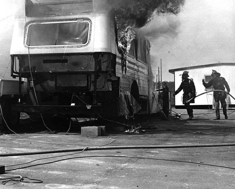 Black and white photo of bus on fire surrounded by a firemen carrying a water hose in 1975. 