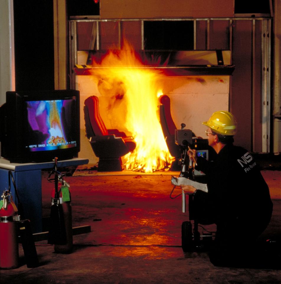 A fire engineer wearing black NIST jumpsuit and yellow hard hat is kneeling next to a tv screen displaying multiple colors of the yellow and orange fire burning on a chair in the background.