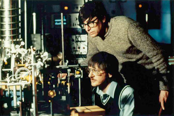 Bill Phillips standing in his lab with postdoc John Prodan seated.