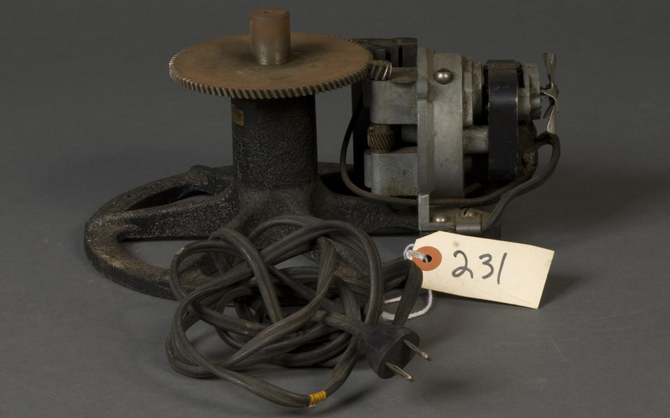 geared mechanism with an electric plug. Attached to the plug's wire is a tag that reads "231." The device is geared to another smaller gear on a device with what looks like a flywheel coming from the front of it. 