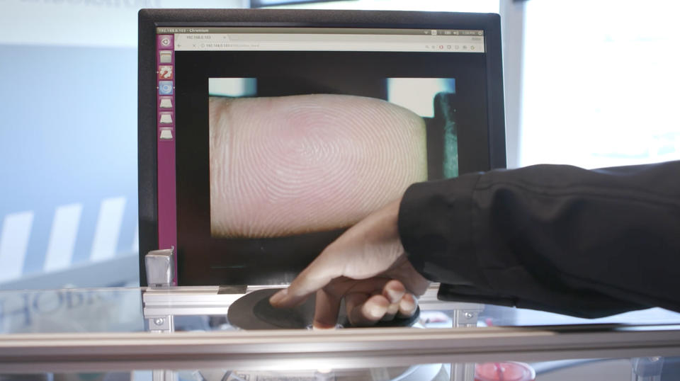 A finger presses on a fingerprint scanner. In the background, the fingerprint is shown on a computer monitor.