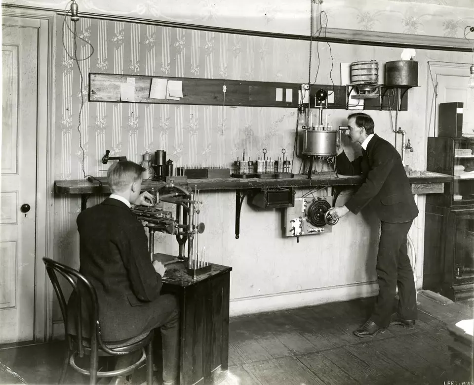 Man sits at a desk on the left while a man on the right stands looking into a device