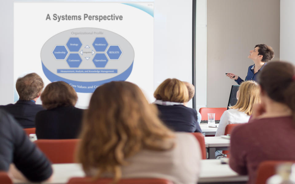 A female Baldrige Ambassador doing a slide presentation of Baldrige to a group of people in a conference room.