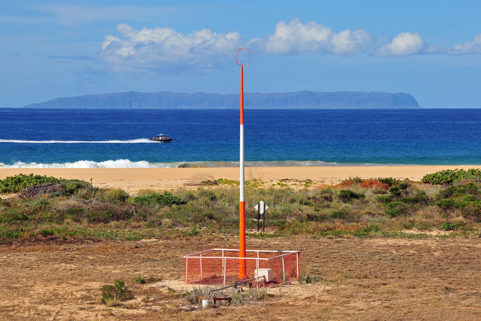 An orange and white antenna a few meters away from a beach. A boat speeds past in the distance. Beyond that the island of Niihau is visible. 