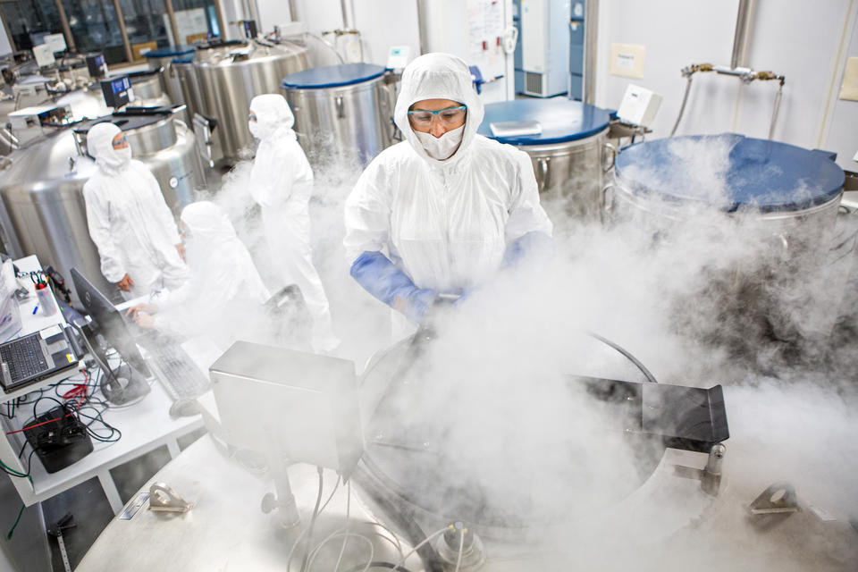 Giant metal tubs are being opened and white vapor is puffing up into the face of one researcher who is in a protective suit with hood, gloves and eye protection. In the background, several other suited researchers discuss something.