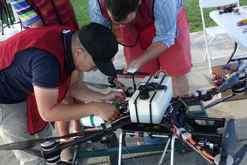 Two men in red smocks make adjustments to their drone