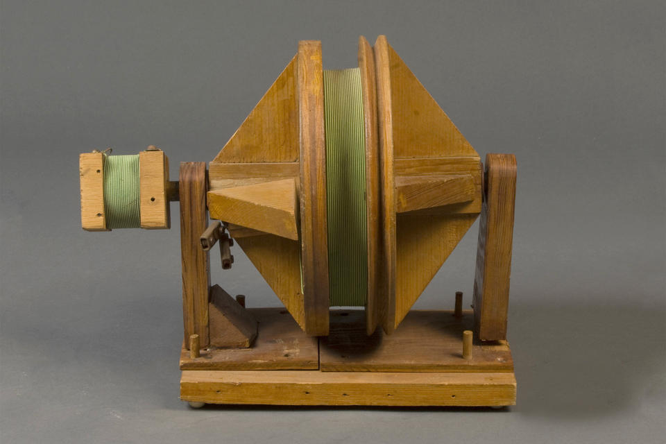 a wooden spindle with green wire wrapped around the middle. to the left of spindle is a smaller block with wire wrapped around it