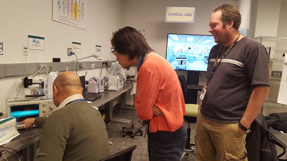 One person sits in front of a wireless infusion pump. Two people stand and look over his shoulder. Other pumps are in the background.