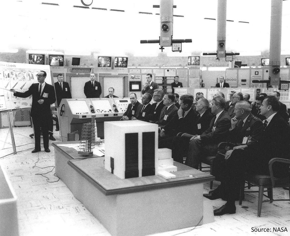 Two men stand in front of poster boards. A bunch of men are seated watching them present.