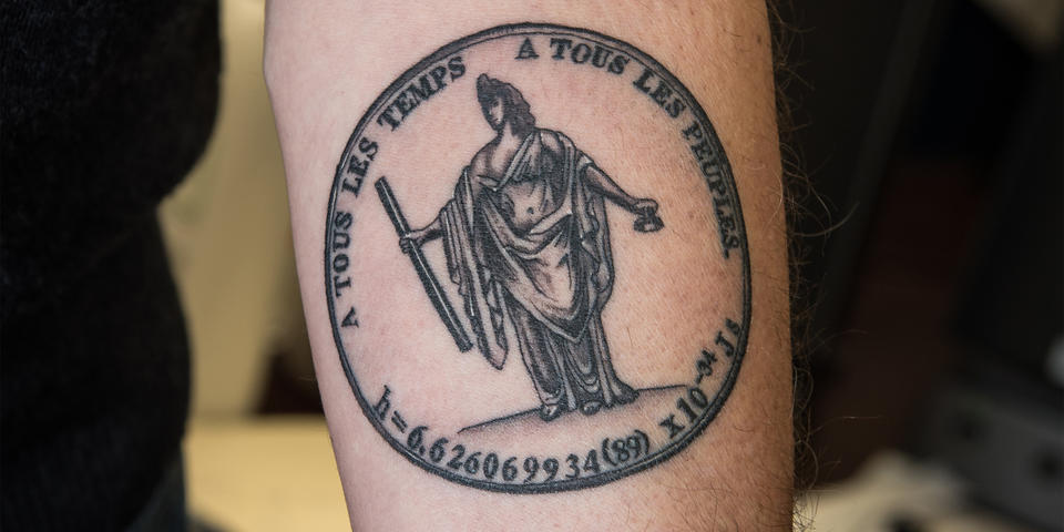 Jon Pratt shows off a tattoo featuring a statue holding a meter bar and a kilogram artifact. The words "For all times, for all peoples" are written in French above, and the final value for the Planck constant is written below.