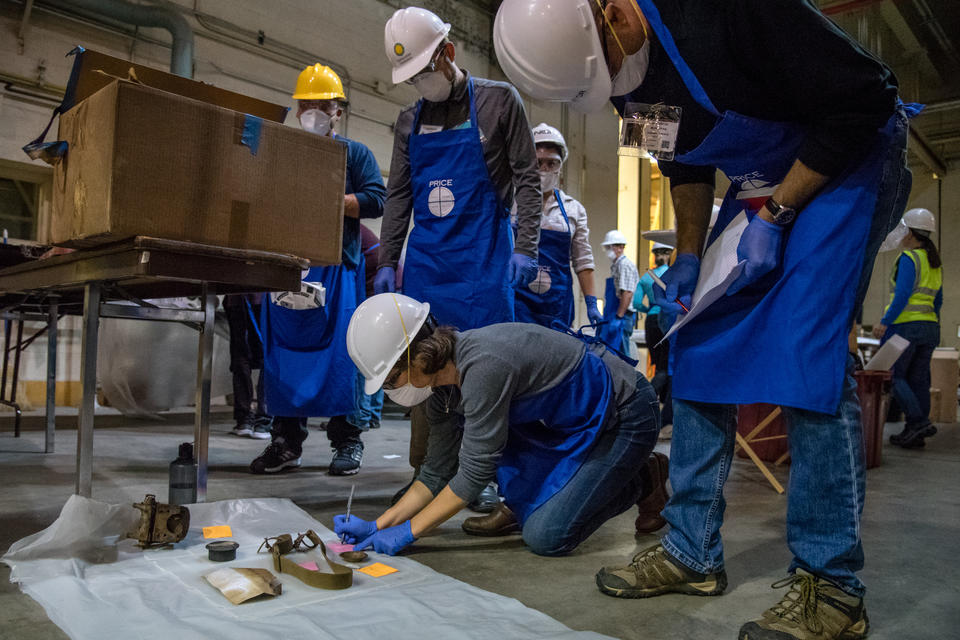 Smithsonian employees wearing hard hats, blue smocks, and respirators kneel over artifacts and record them on their clipboards