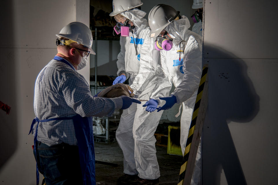 People wearing full body white hazard suits, hard hats, safety glasses and face masks emerge from burn test room with clay mask artifact