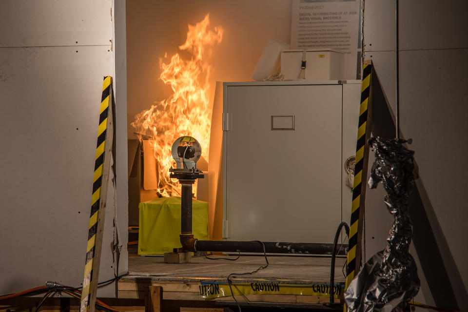 view through the doorway of the simulated storage room. The fire grows as a video camera in the room looks on. 