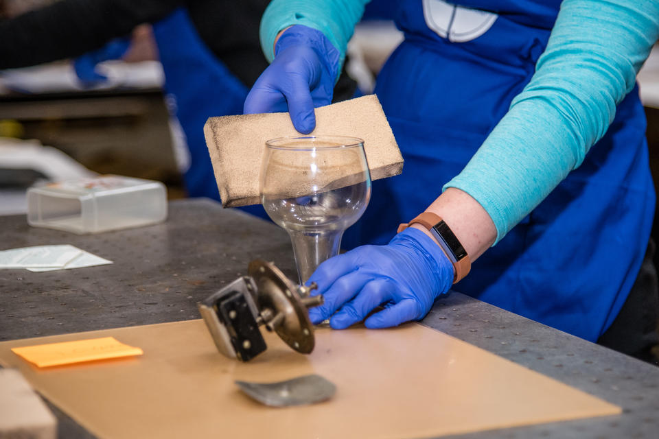 Smithsonian curator wearing blue gloves and a blue smock carefully wipes away soot from a glass goblet with a sponge
