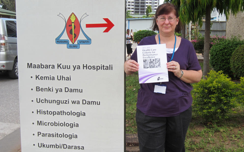 Pat Griffith, a Baldrige examiner, is holding the Baldrige Excellence Framework booklet in Tanzania next to a hospital sign in Swahili.  