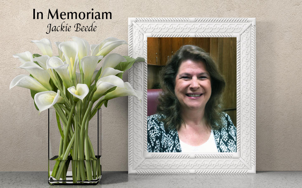 In Memoriam: Photo of Jackie Beede with white lilies in a vase beside frame.