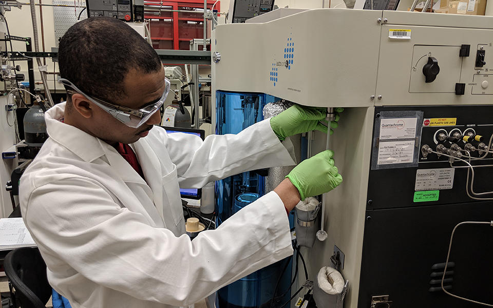 man in a white lab coat and safety goggles loads a metal oxide framework material into a testing machine