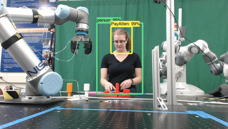 Researcher moves differently shaped and colored plastic blocks on a table as a robotic arm looks on