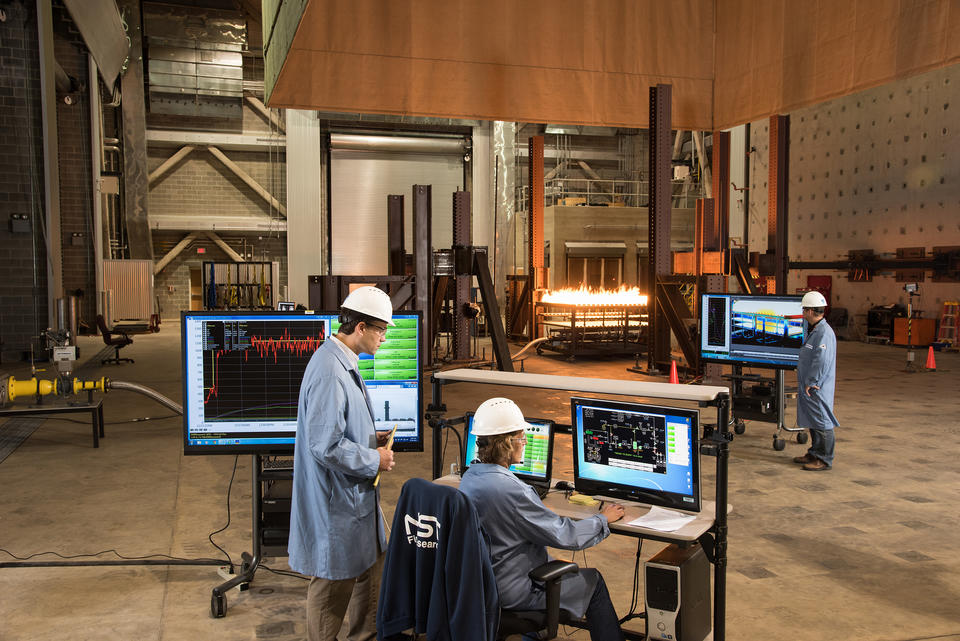 Three individuals watch measurements on a screen while a test fire burns