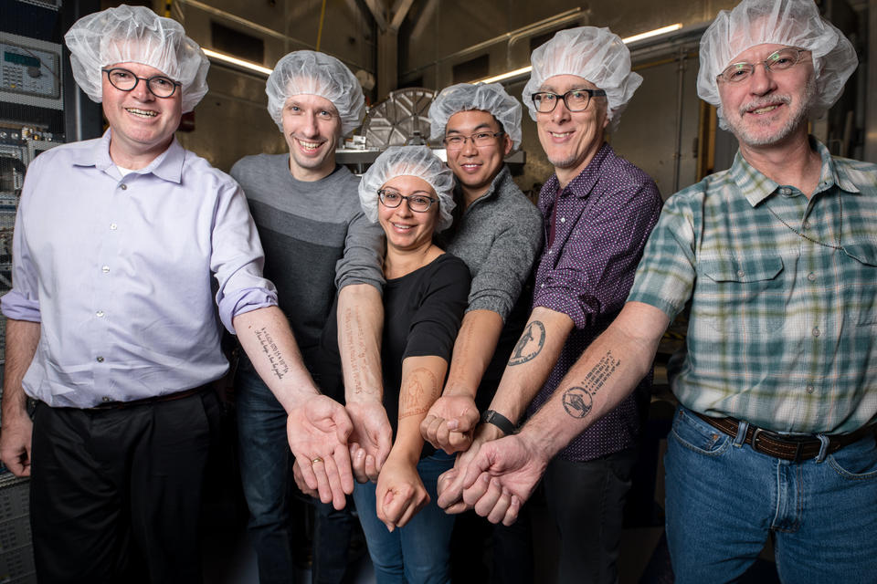 NIST researchers wearing hairnets proudly and happily present their forearms bearing tattoos of the value of the Planck constant