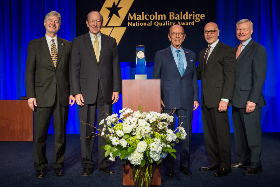 Center: Baldrige Award on a podium. Left: 2 men in suits. Right: 3 men in suites