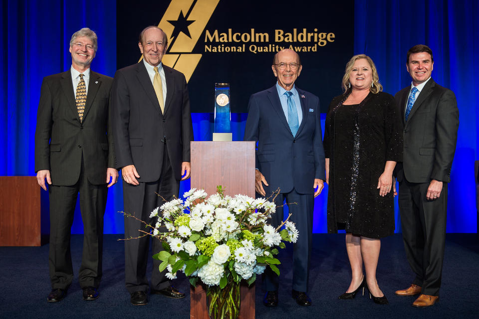 Center: Baldrige Award on a podium. Left: 2 men in suits. Right: man in suit, woman in dress, man in suit