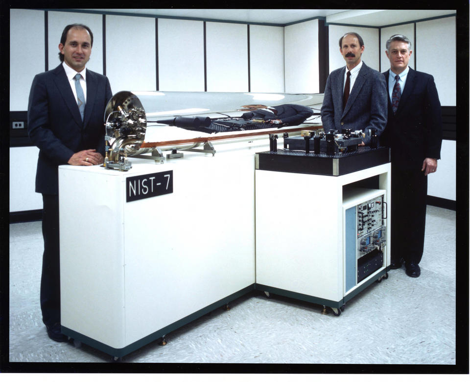 Three men stand behind a glass tube with equipment inside