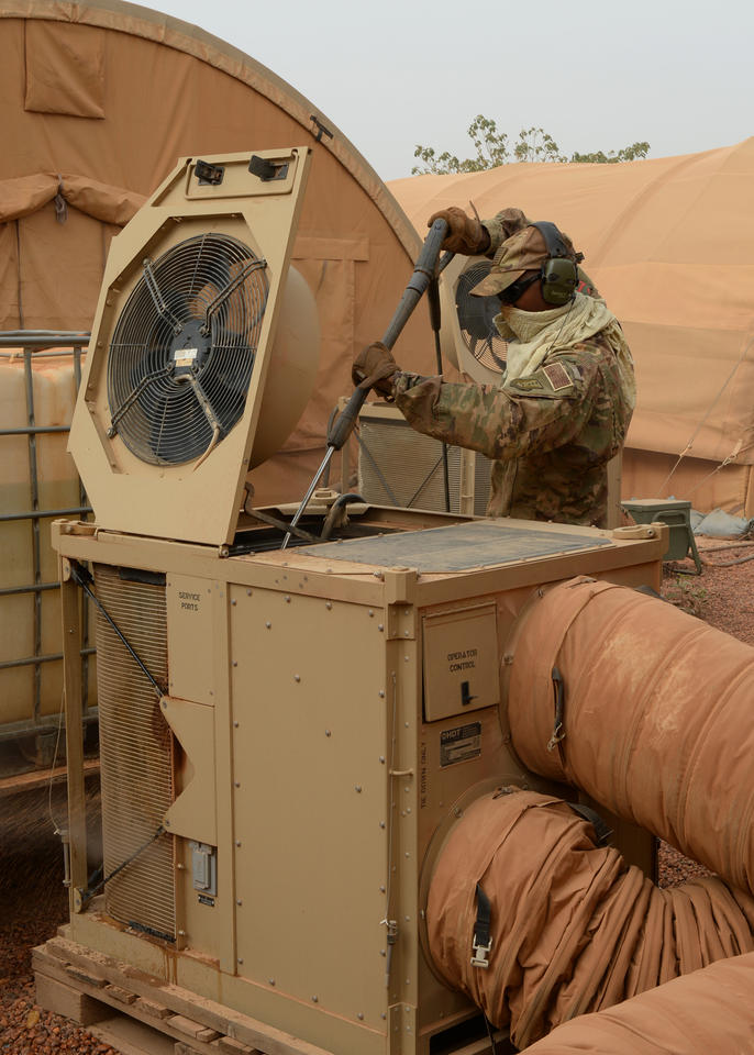A man in brown camouflage clothing, including baseball cap, gloves and ear protection, uses a long pole to clean the inside of an HVAC unit outside a military tent.