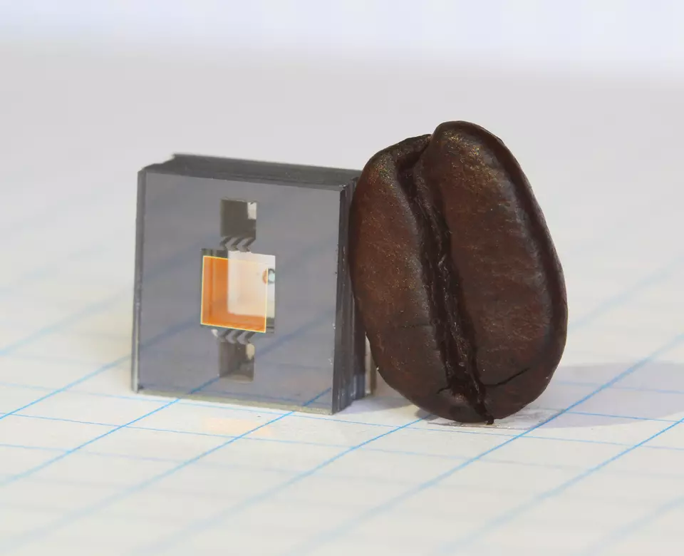 Square gray cube with two square holes in it and a gold square in between the holes, and a dark brown coffee bean leaning against the cube.