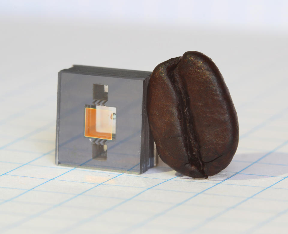 Square gray cube with two square holes in it and a gold square in between the holes, and a dark brown coffee bean leaning against the cube.