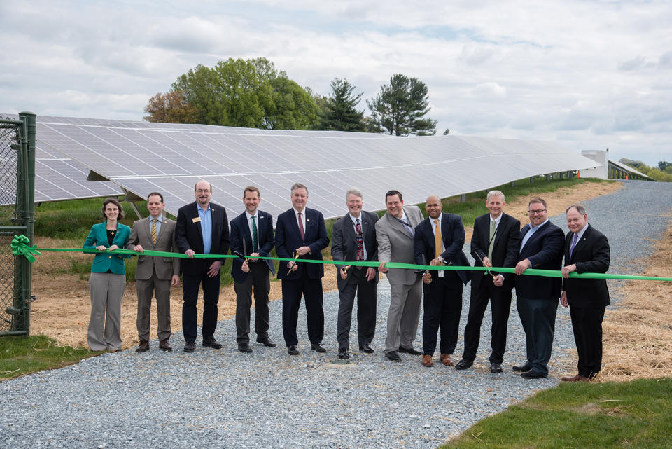 Eleven people stand in front of the NIST solar array getting ready to cut an orange ribbon with large scissors.