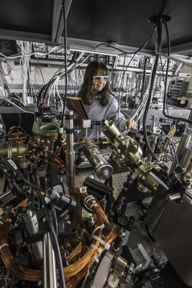 woman in goggles standing behind a lab table filled with equipment and wires. She has a lab notebook in her hand.