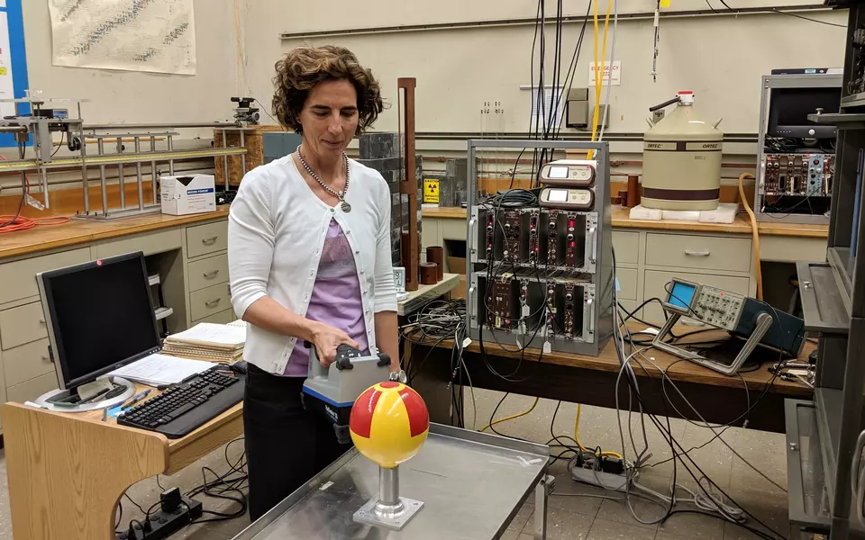 Woman standing in a lab holds a rectangular radiation detector near a metal globe that has been painted with a radiation symbol
