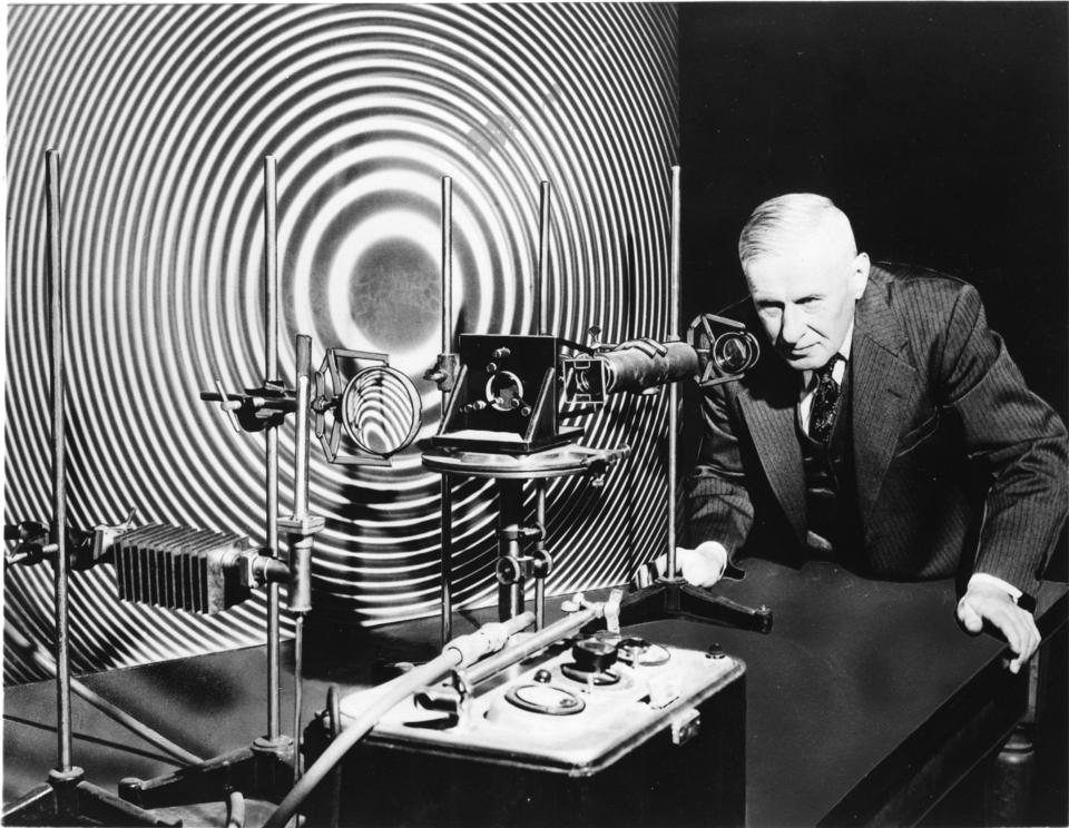Man stands looking into the eyepiece of scientific equipment. Pattern behind is black and white concentric rings.