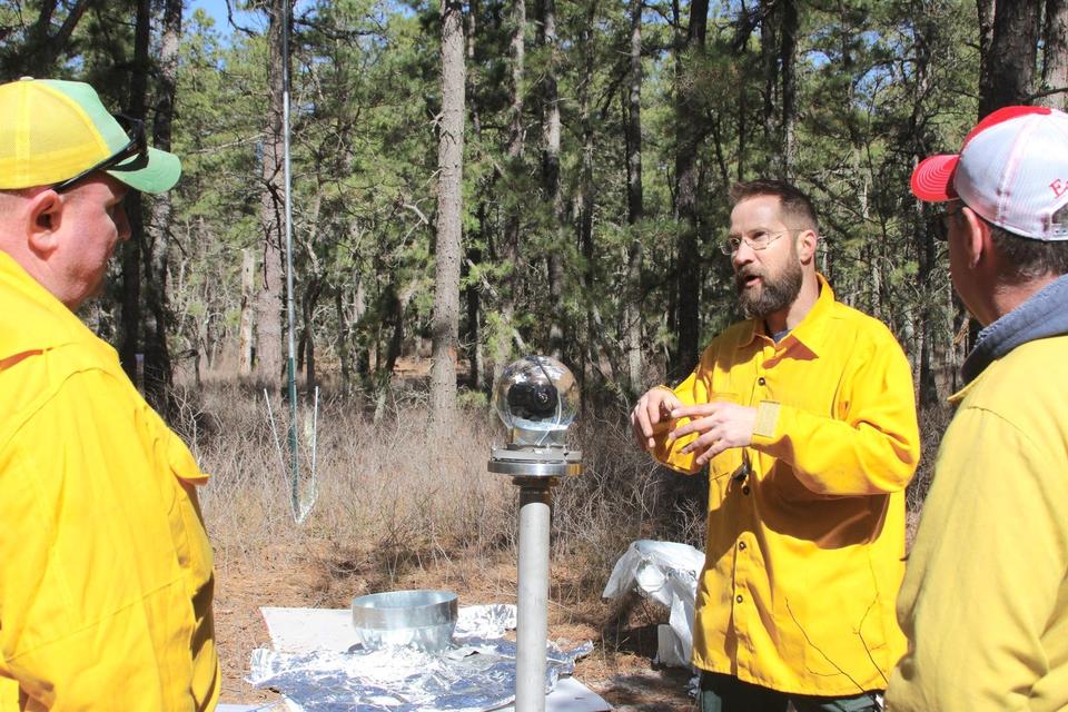 NIST engineer in yellow jacket stands to right of BOB glass globe. To his right and left are two men.