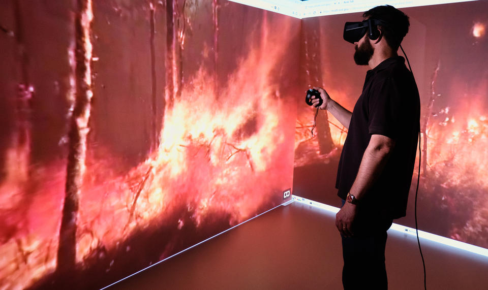 A profile of a man wearing VR equipment is seen against two large screens displaying the view from within a forest fire.
