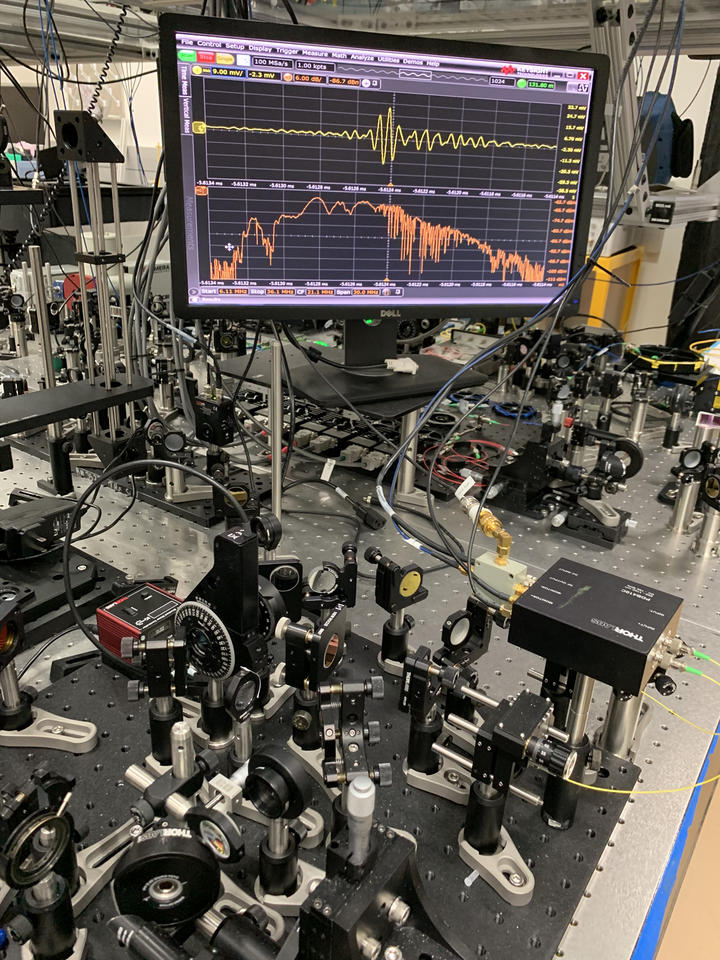 Table covered with lenses and other optical equipment, and a flat screen mounted high in the background showing a yellow squiggly line across the top and a spiky orange line below.