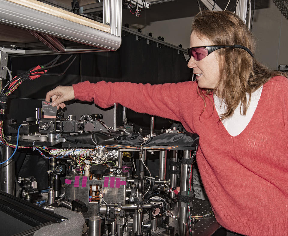 Woman with reddish hair and wearing a pink sweater and laser safety glasses reaching over an instrument table and holding a card used to path of lasers.