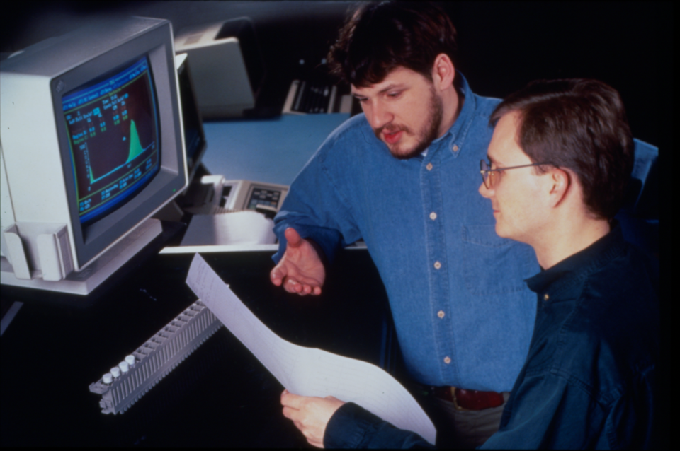 Two men look at a printout in front of a computer screen