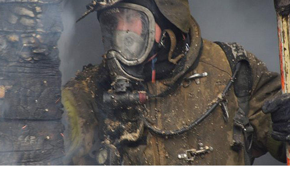 Firefighter wearing gear, including face shield, standing near smoking building