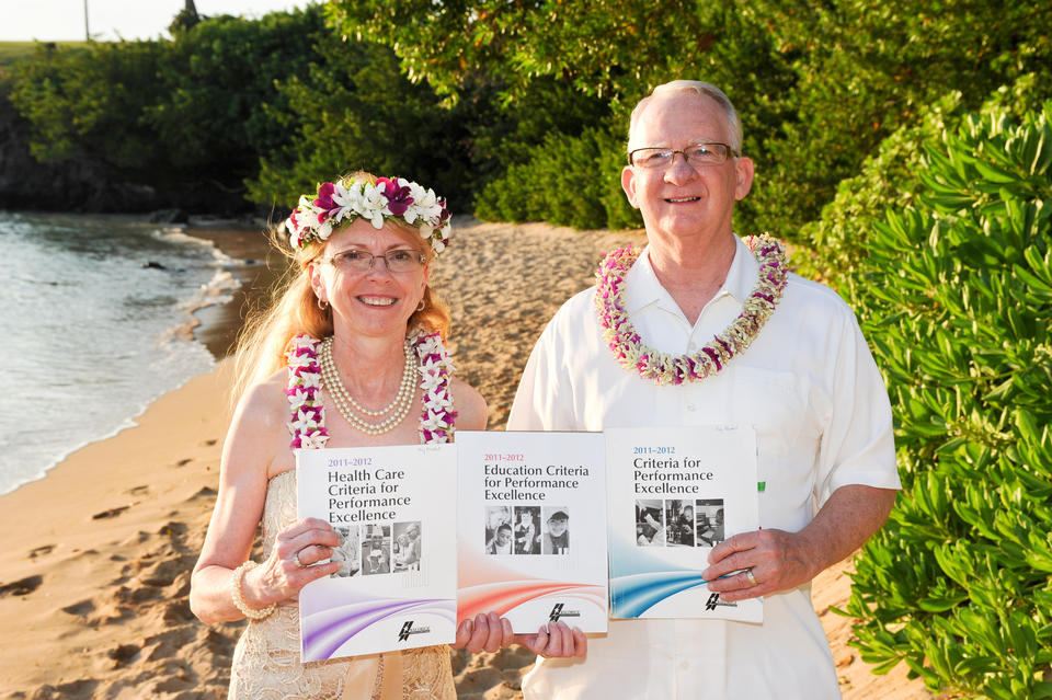 Newly wed examiners Kay Kendall and Glenn Bodinson pose together in Hawaiian dress holding Baldrige booklets on a tropical beach