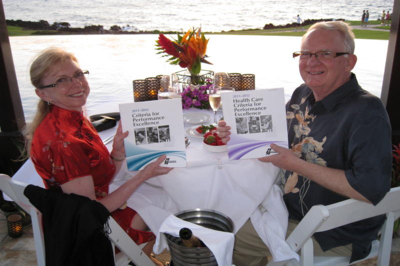 Newly examiners Kay Kendall and Glenn Bodinson pose with Baldrige booklets while dining in a tropical setting
