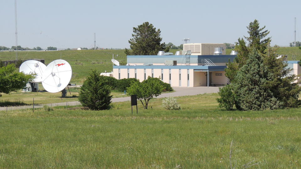 a building in a field with a few trees around it, two big radio dishes at left and some tall radio transmission antennae in the background