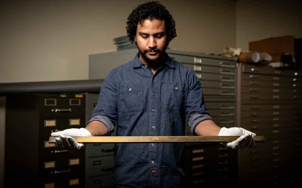 Man wearing white gloves holding a rectangular brass bar out in front of him.