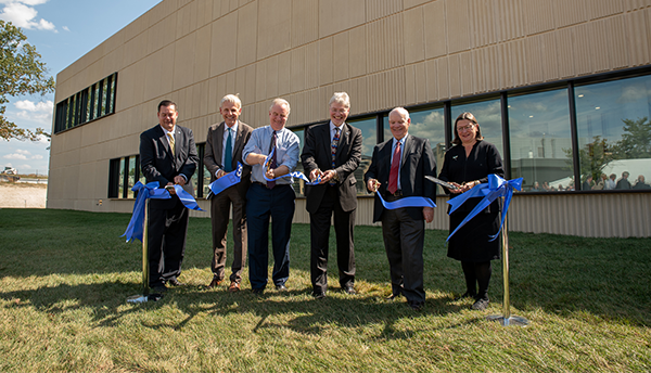 Six people stand in front of a large tan building, cutting a blue ribbon.